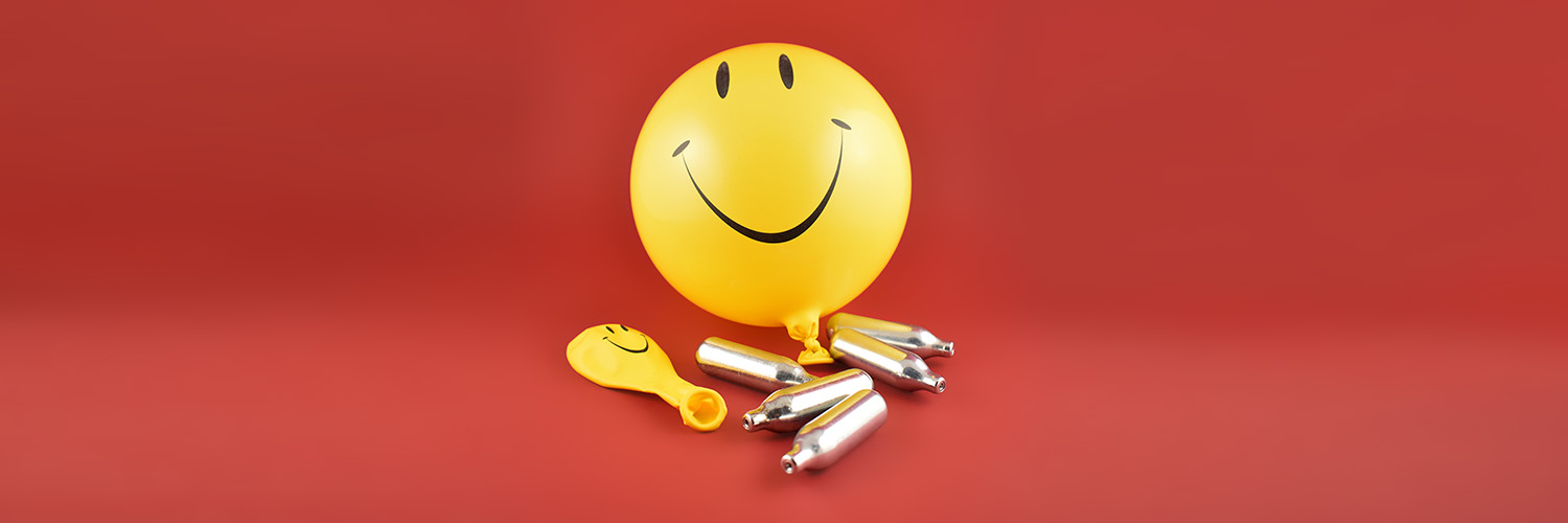 Lachgasflaschen mit gelbem Smileyballon vor rotem Hintergrund