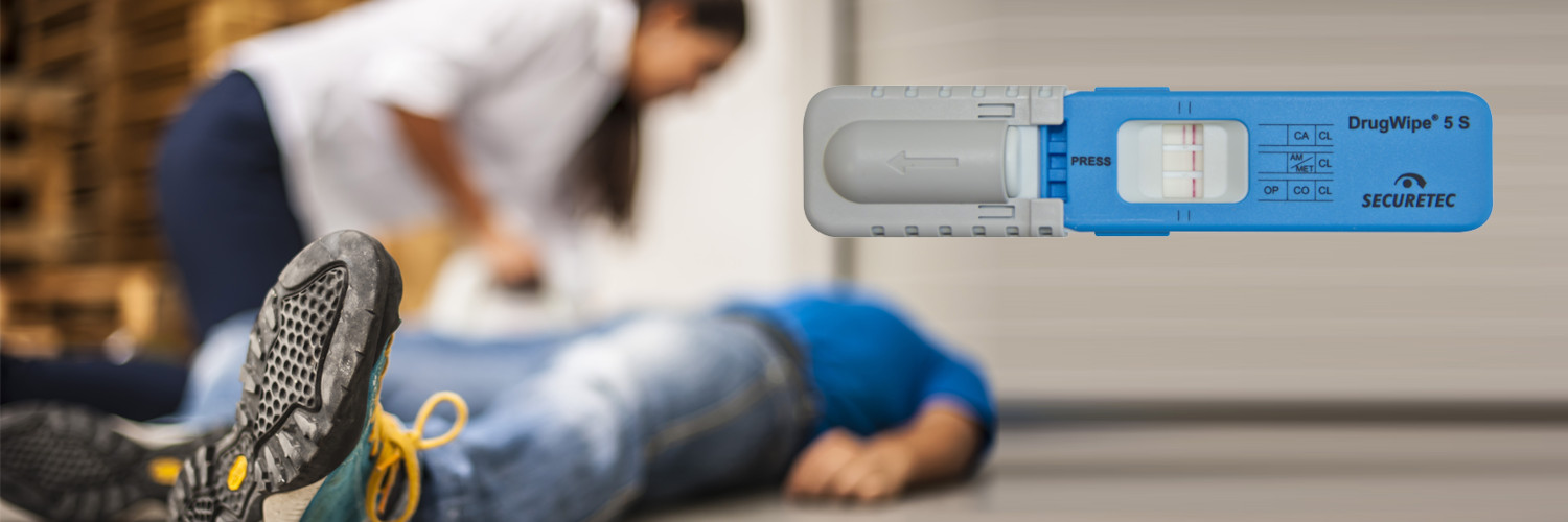 Eine Mann liegt ohnmächtig am Boden und eine Kollegin spricht ihn an. Im Vordergrund ist der Drogenschnelltest DrugWipe zu sehen.