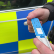 Blau-weisser Drogenschnelltest DrugWipe wird vor einem Polizeiauto mit zwei Händen gehalten